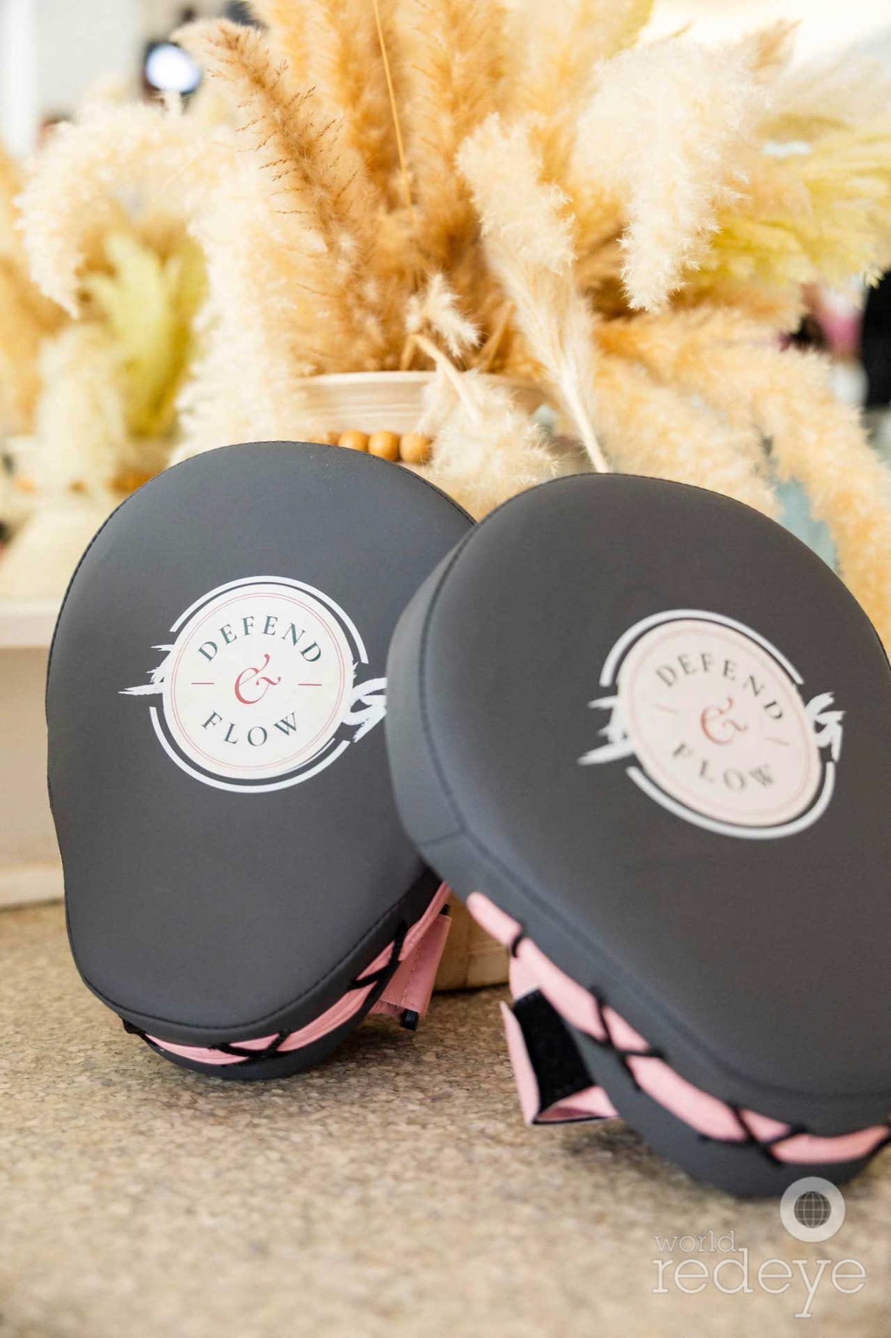 Two pink and black Defend & Flow focus mitts rest on a surface beside pampas grass.
