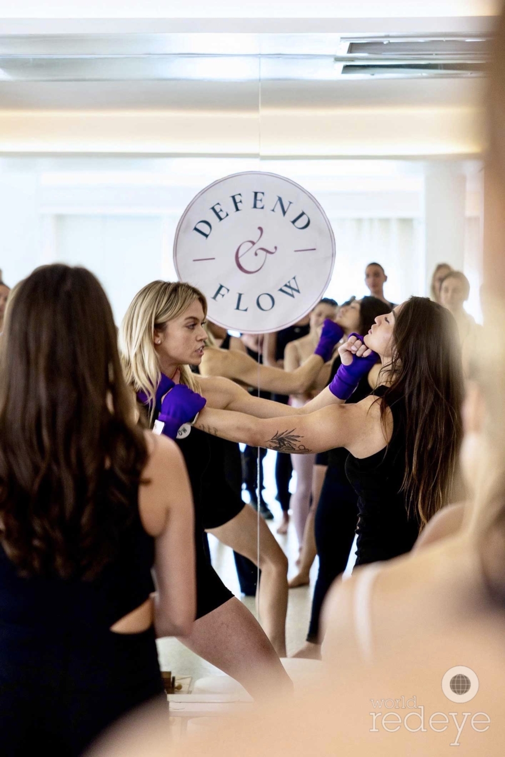 Two women practice a self-defense technique reflected in a studio mirror.