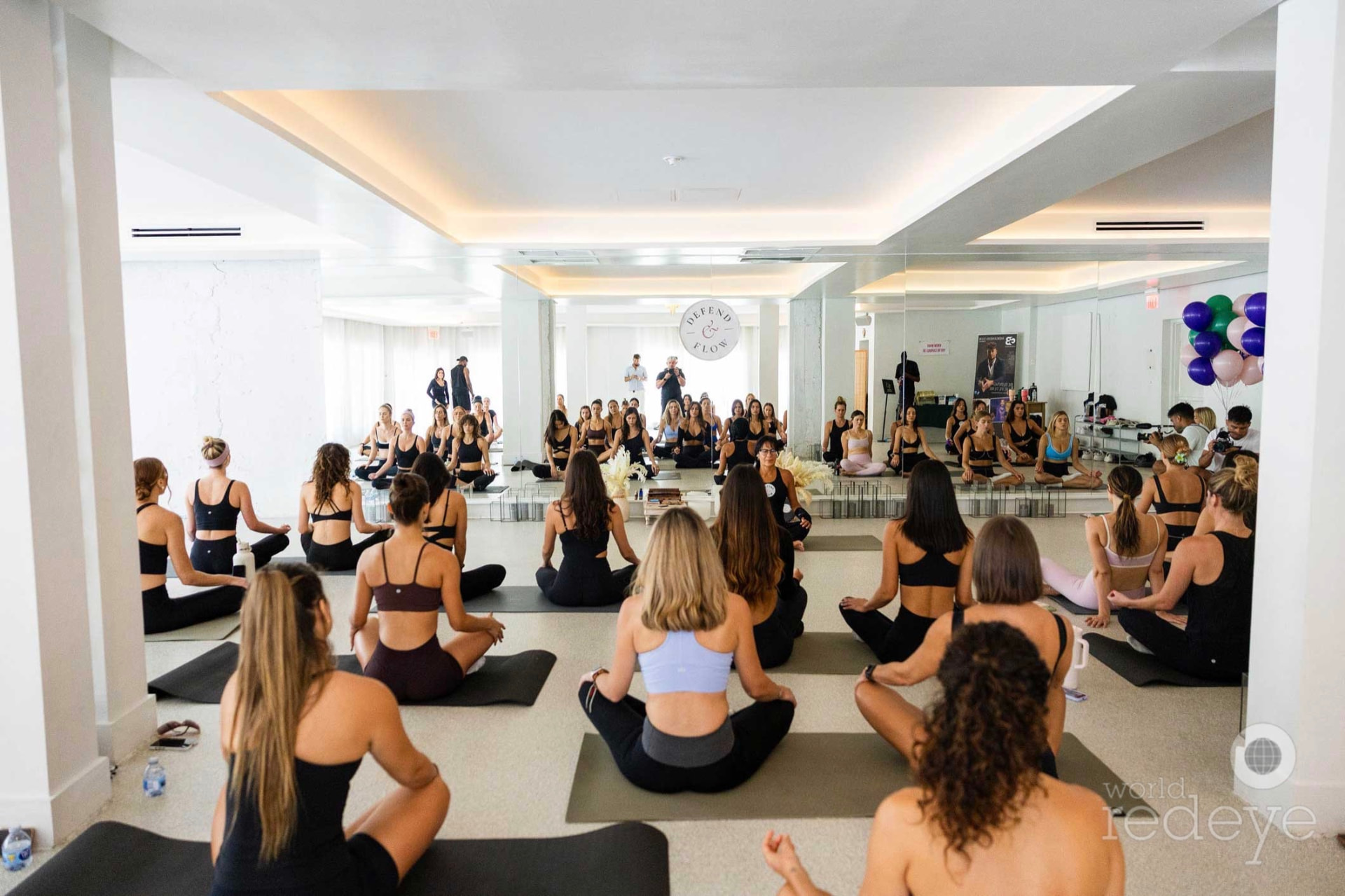 A large group of women in black activewear sit in meditation on yoga mats in the mirrored studio.