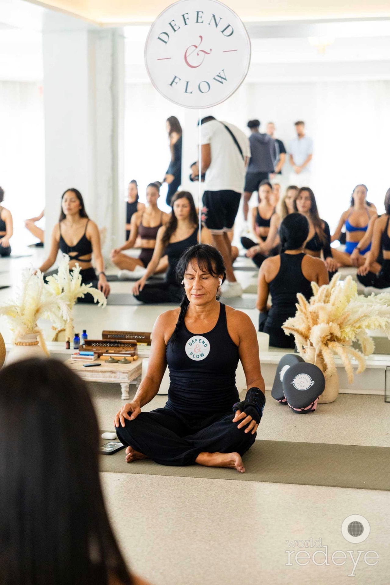 A woman wearing a Defend & Flow badge leads the group in a seated meditation.